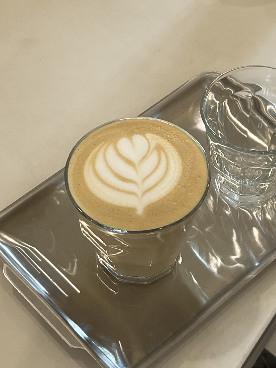 A latte with a leaf-shaped latte art, served with a glass of water on a metal tray